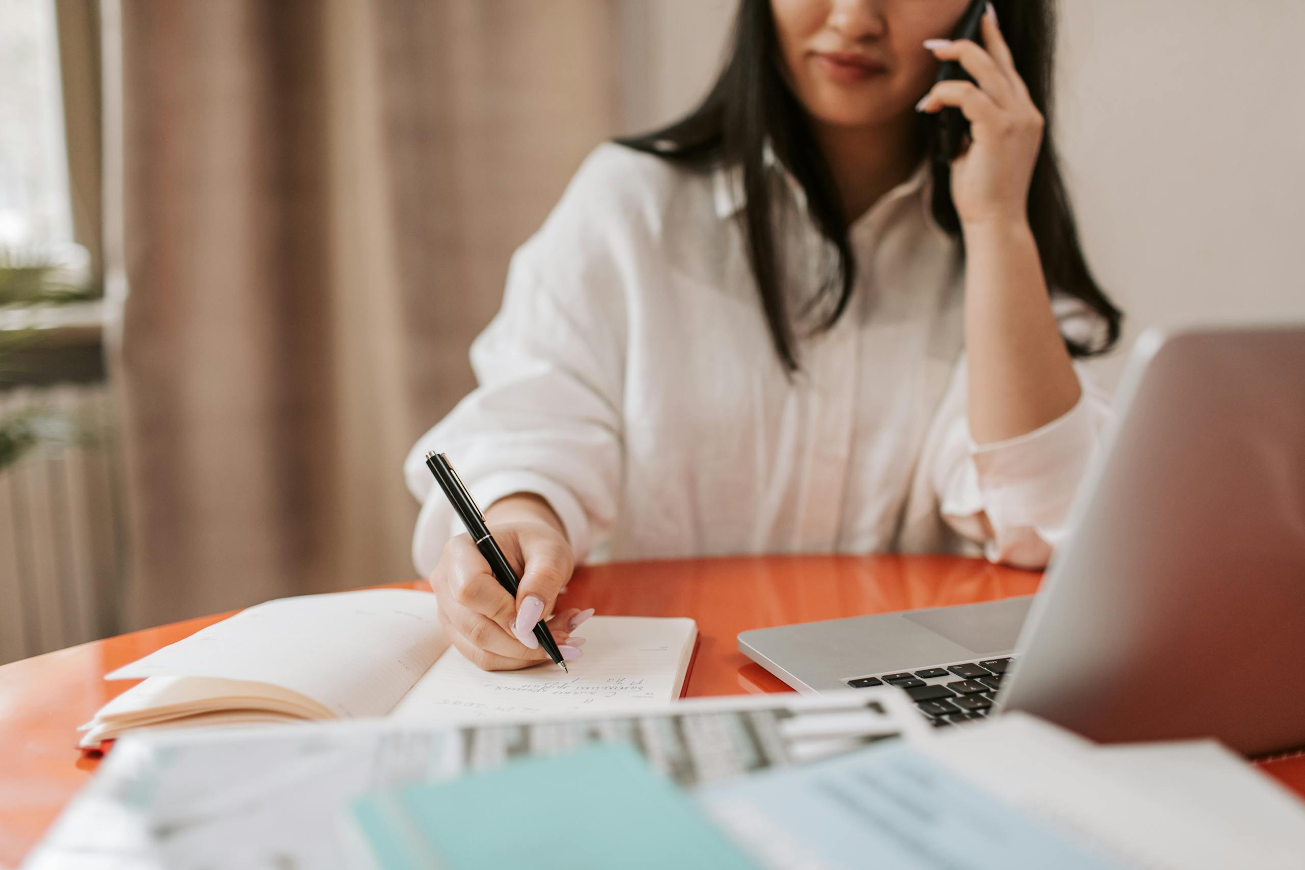 Woman multitasking by writing notes while talking on the phone, seated at a desk with a laptop.